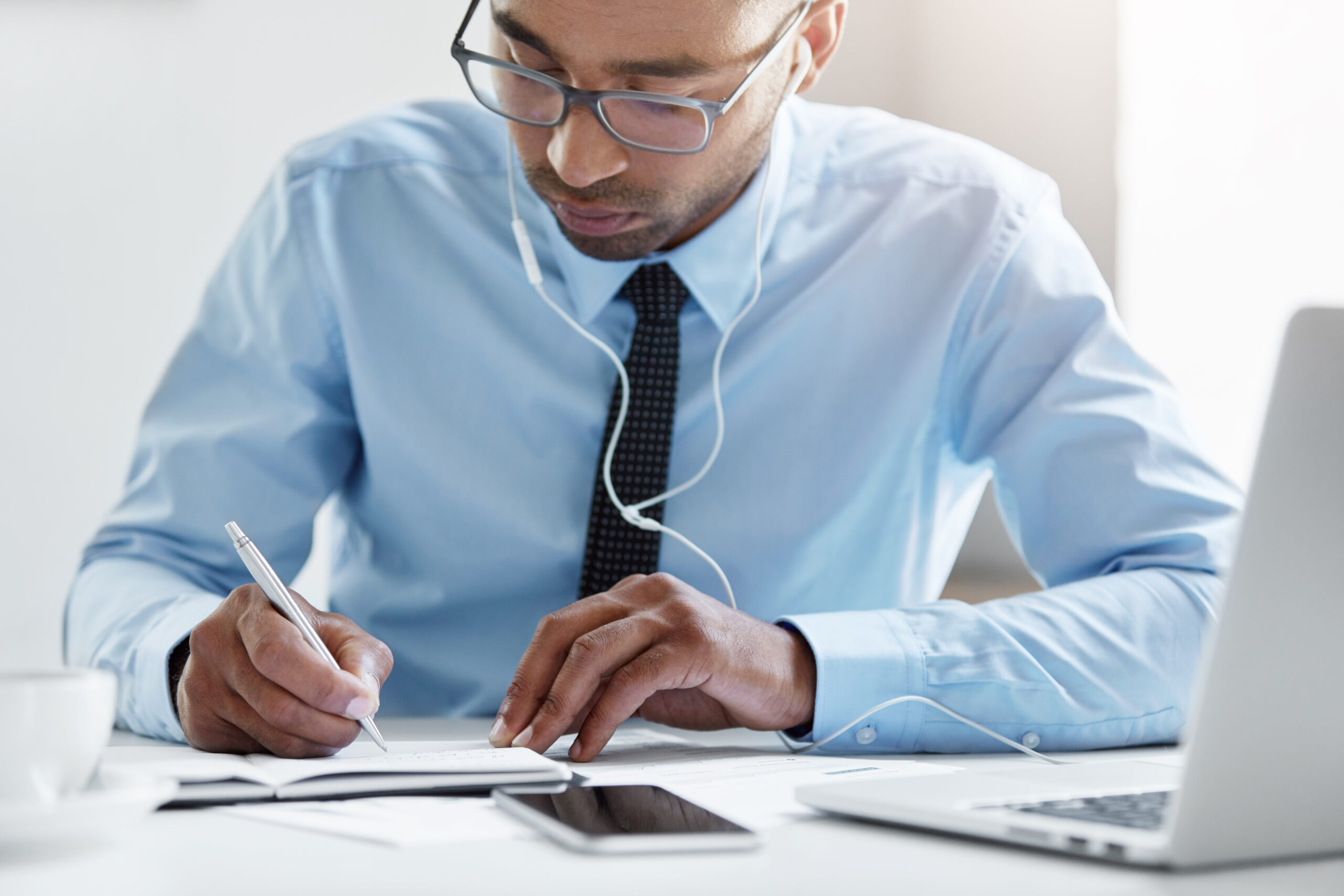 Photo of young financier with serious look dressed in formal clothes, making annual financial report and listening audio book with earphones using laptop computer. Technology and business concept